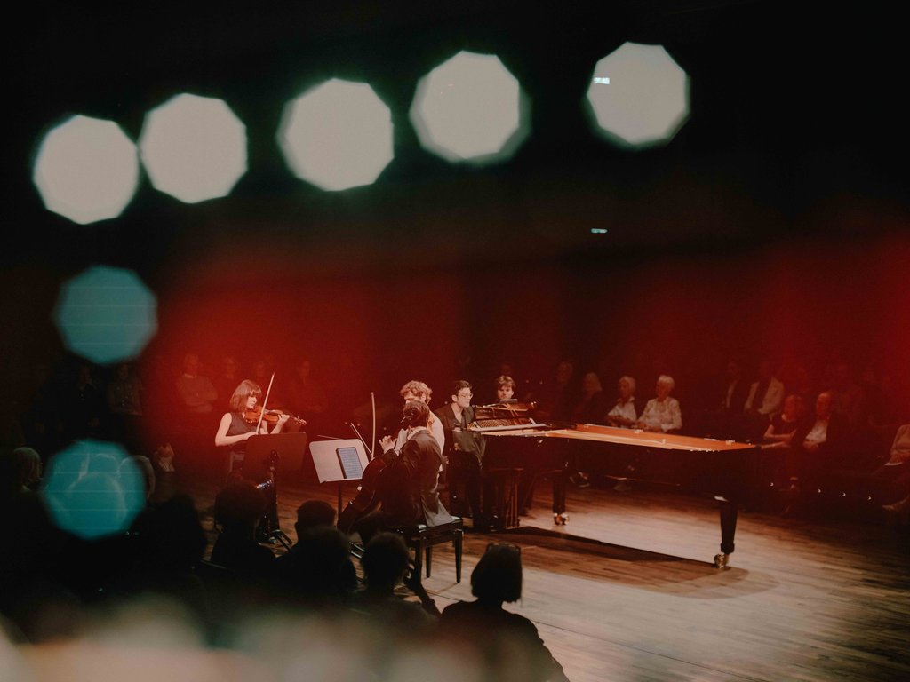 Musiker spielen klassische Musik auf einer Bühne. Ein Violine und ein Klavier sind sichtbar, das Publikum sitzt im Hintergrund.