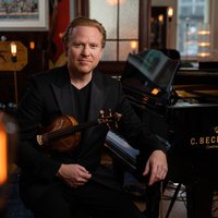 A man in a suit sits next to a C. Bechstein piano, holding a violin in his hand.