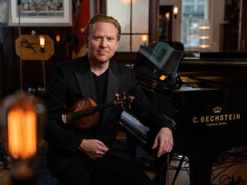 Ein Mann in einem Anzug sitzt neben einem C. Bechstein-Flügel. Er hält eine Geige in der Hand.