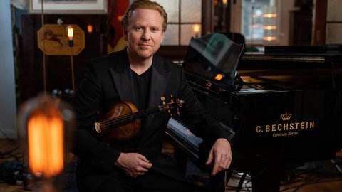 A man in a suit sits next to a C. Bechstein piano, holding a violin in his hand.