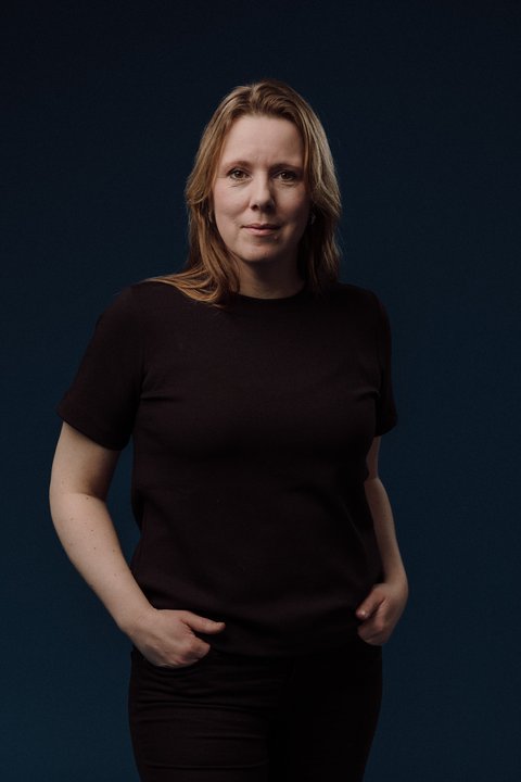 Ein stehendes Porträt einer Frau mit halblangen blonden Haaren. Sie hat ihre Hände in die Hosentaschen gesteckt und lächelt entspannt. Sie trägt schwarze Kleidung. Der Hintergrund ist schwarz. © Marius Fiskum