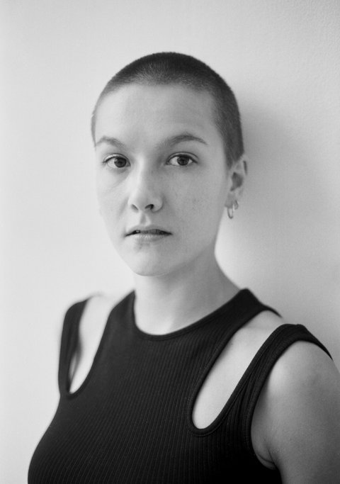 Eine Person mit kurzrasierten Haaren posiert vor einer hellen Wand und blickt in die Kamera.
