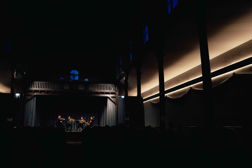 A small ensemble performs on stage in a dimly lit hall with classical architectural style.