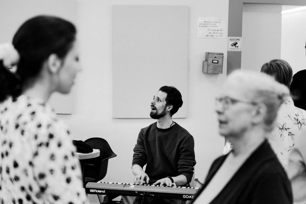 Black and white photo: A man plays the keyboard in a room with singing people, some blurred in the foreground.
