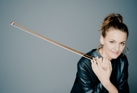 A woman holding a violin bow smiles at the camera. She is wearing dark clothing and her hair is tied back.