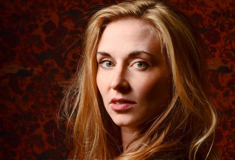 Eine Frau mit langen, blonden Haaren posiert für ein Portraitbild vor einem rot-schwarzen Hintergrund. Sie trägt einen schwarzen Pullover und blickt über ihre Schulter in die Kamera.