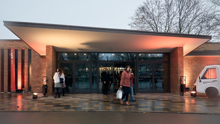 People entering a modern building with a glass facade, illuminated in the evening for an event. Trees are visible in the background.
