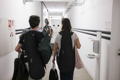Zwei Personen gehen einen weißen Flur entlang. Sie tragen Instrumentenkoffer, wahrscheinlich für Geigen oder andere Saiteninstrumente.