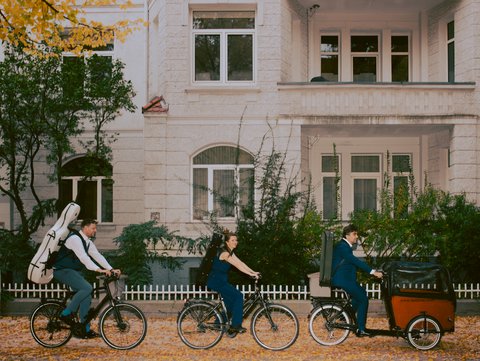 Drei Menschen fahren mit Fahrrädern durch eine herbstlich bepflanzte Straße. Einer hat ein Cello, eine eine Violine und einer eine Viola auf dem Rücken. Reihenhäuser im Hintergrund.
