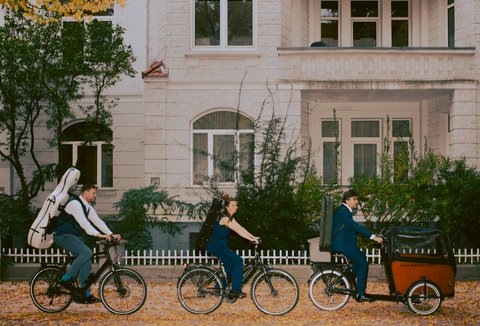 Drei Menschen fahren mit Fahrrädern durch eine herbstlich bepflanzte Straße. Einer hat ein Cello, eine eine Violine und einer eine Viola auf dem Rücken. Reihenhäuser im Hintergrund.