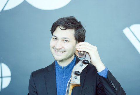 Ein Mann im Anzug posiert mit einem Violoncello und lächelt.