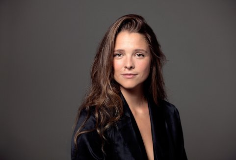 A woman is standing in front of a dark background. She is wearing a dark blouse. She is looking into the camera.