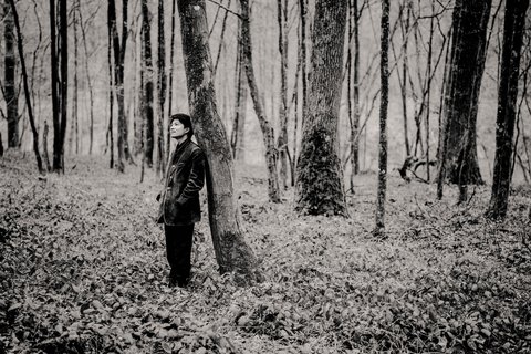 Eine Person steht in einem Wald, lehnt an einem Baum. Die Szene ist in Schwarz-Weiß gehalten, wirkt ruhig und nachdenklich.