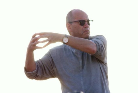 A man is standing in front of a bright background. He is looking away from the camera and posing with his arms raised.
