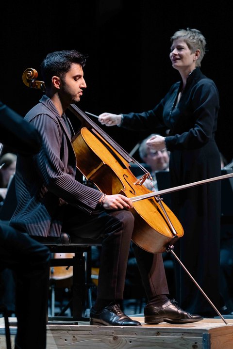 Ein Cellist sitzt auf der Bühne und spielt sein Instrument, während eine Dirigentin das Orchester leitet.