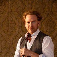 A man with curly hair in a white shirt and grey vest holds a violin against a decorative background.