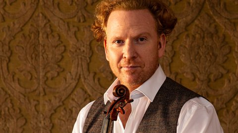 A man with curly hair in a white shirt and grey vest holds a violin against a decorative background.
