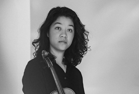 Black and white image of a woman holding a violin. She stands against a light background, wearing a dark outfit.