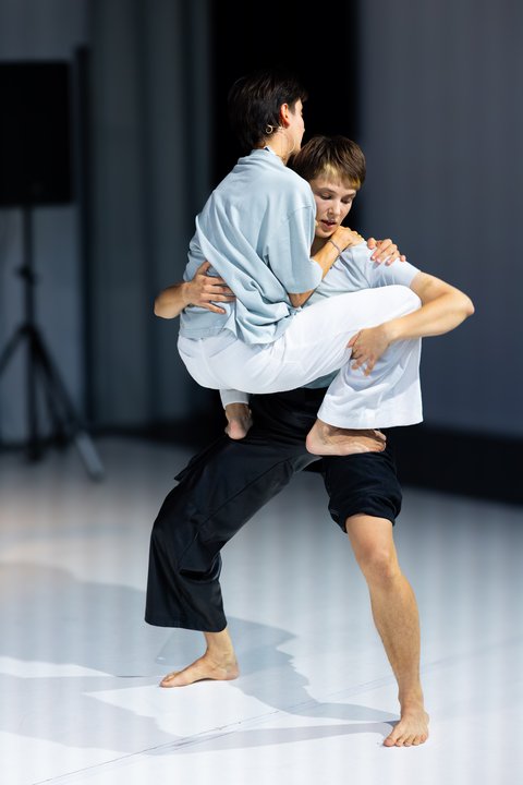 Two dancers on stage, one lifting the other in a dynamic pose. They are barefoot and the stage is minimalistic.