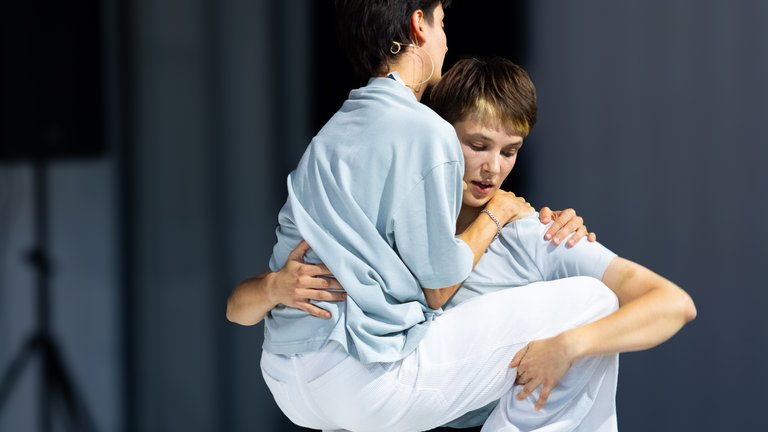 Two dancers on stage, one lifting the other in a dynamic pose. They are barefoot and the stage is minimalistic.