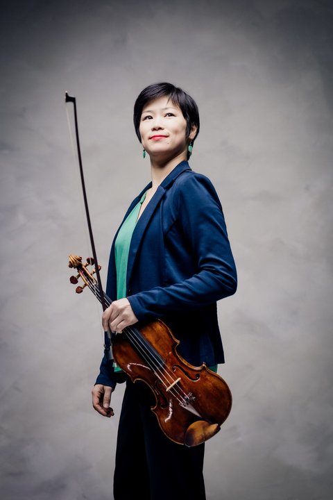 Eine Frau mit einer Violine steht vor einem hellen Hintergrund. Sie trägt einen Anzug und lächelt. © Zuzanna Specjal