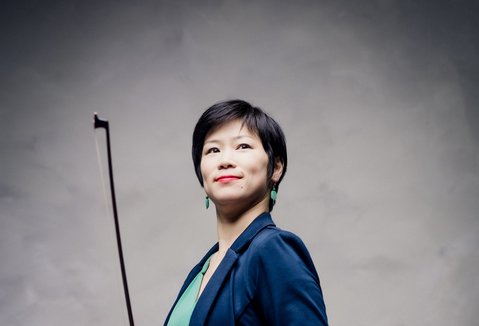 A woman with a violin is standing in front of a light background. She is wearing a suit and smiling.