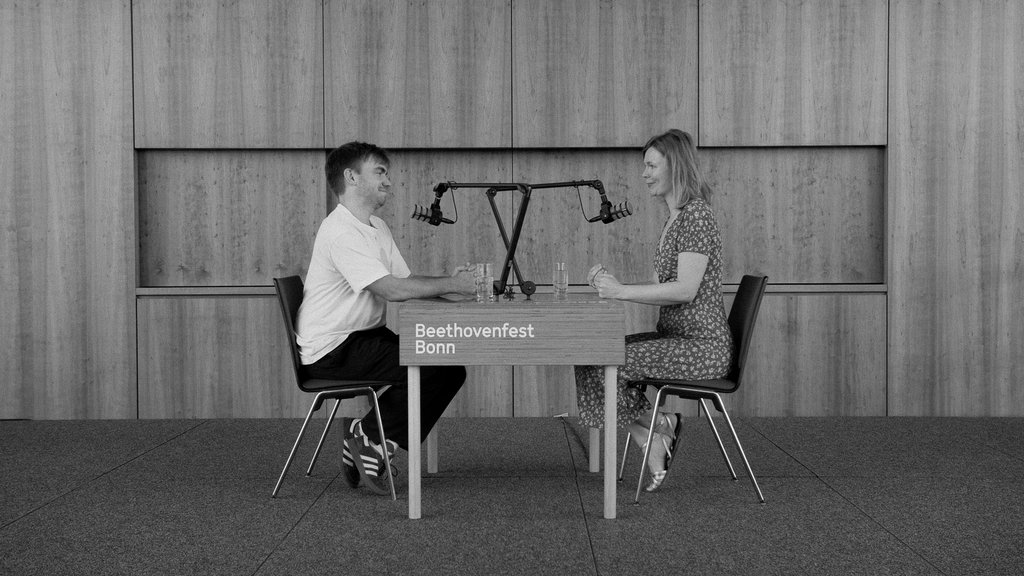 Eine Frau und ein Mann sitzen sich an einem Tisch in einem Saal gegenüber. Sie nehmen einen Podcast auf.