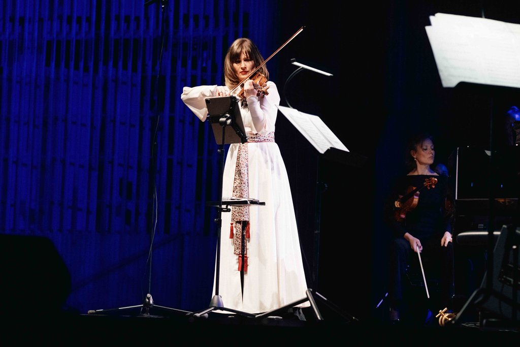 Eine Frau in einem langen weißen Kleid spielt Geige auf der Bühne. Im Hintergrund sind weitere Musiker zu sehen.