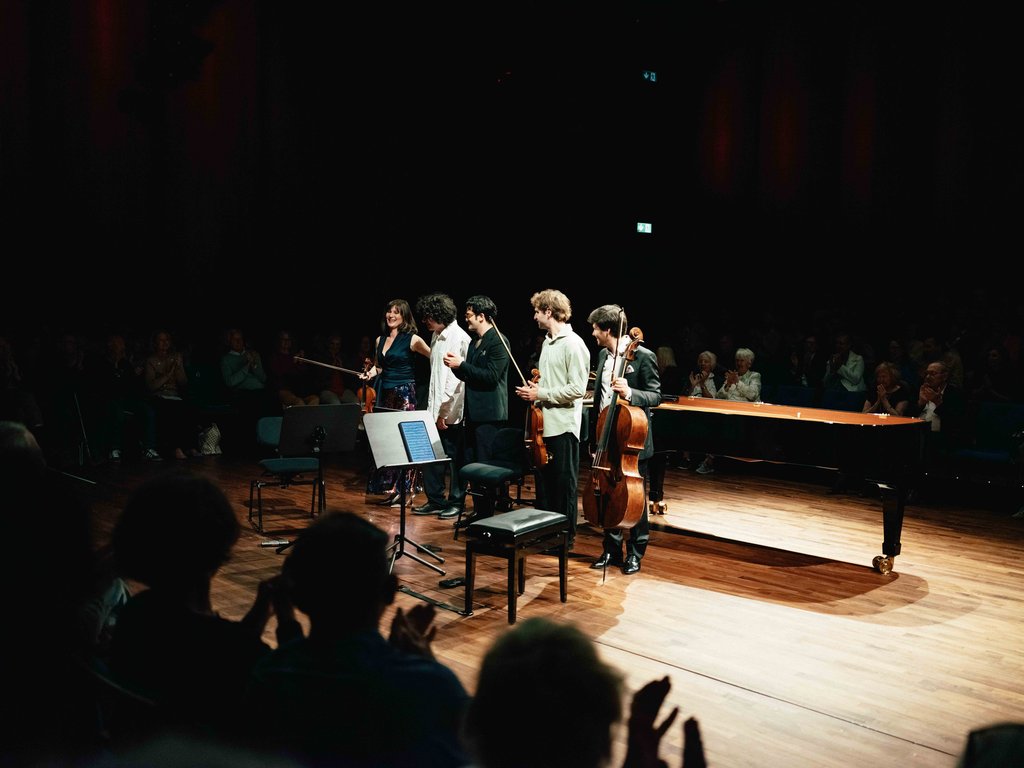 Ein Quartett verbeugt sich auf einer Bühne vor einem applaudierenden Publikum. Ein Cello und ein Klavier sind sichtbar.
