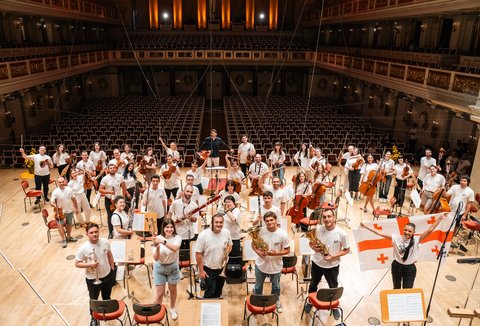 Eine große Gruppe von Orchestermusikern steht auf einer Bühne. Sie tragen weiße Hemden und halten verschiedene Instrumente.
