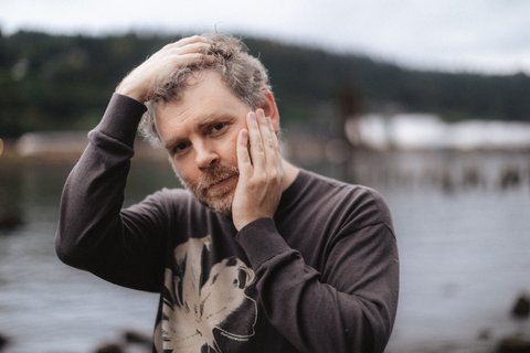 Ein Mann in einem grauen Hoodie posiert vor einem See. Er hält seinen Kopf in seinen Händen und blickt ernsthaft in die Kamera.