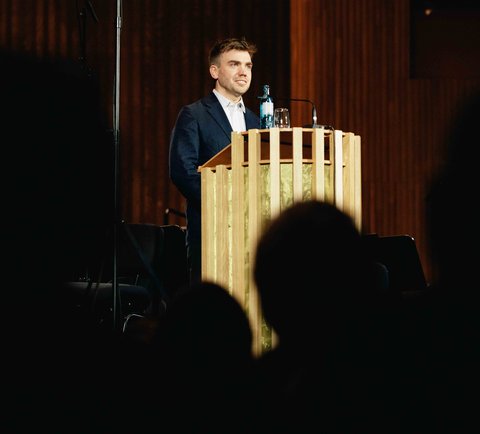 Ein Mann steht an einem Rednerpult vor einem Publikum. Der Raum ist holzgetäfelt und die Beleuchtung ist warm.