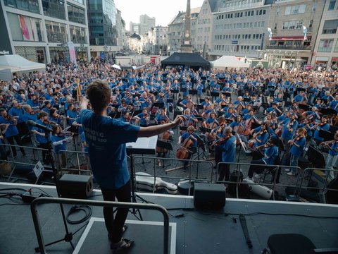 Ein Dirigent leitet ein großes Orchester und Publikum in blau gekleidet auf einem Stadtplatz, umgeben von modernen Gebäuden.