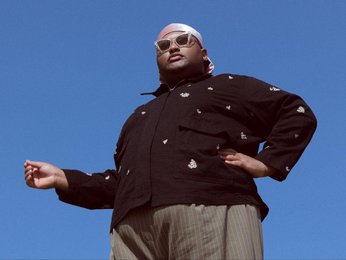 Ein Mann steht vor blauem Himmel, von unten fotografiert, mit einem dunklen Jacke, einem weißen Kopftuch und einer Sonnenbrille, eine Hand ausgestreckt.