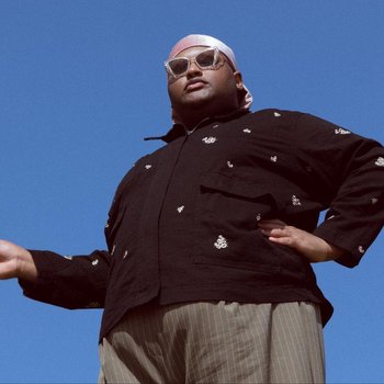 Ein Mann steht vor blauem Himmel, von unten fotografiert, mit einem dunklen Jacke, einem weißen Kopftuch und einer Sonnenbrille, eine Hand ausgestreckt.