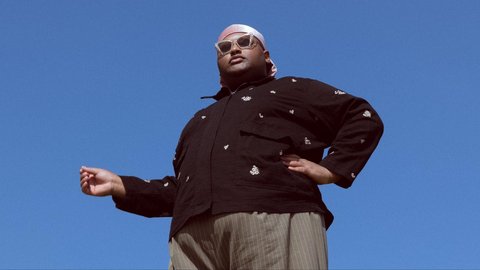 A man stands against a blue sky, photographed from below, wearing a dark jacket, a white headscarf and sunglasses, with one hand outstretched.  © Adam Rom