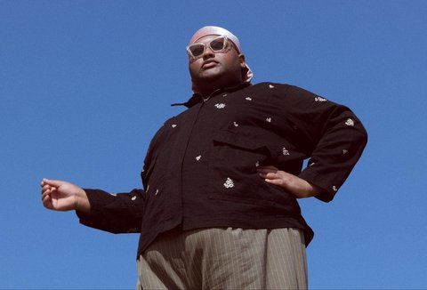 A man stands against a blue sky, photographed from below, wearing a dark jacket, a white headscarf and sunglasses, with one hand outstretched.