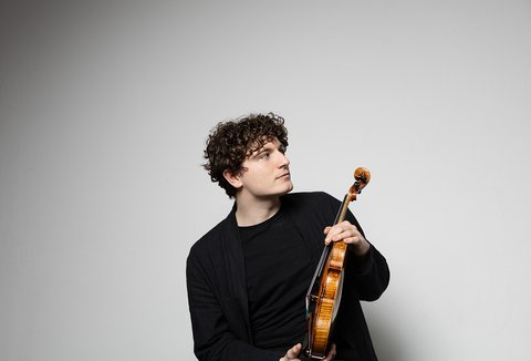 A young man with curly hair holds a violin in his hands, looking thoughtfully to the side.