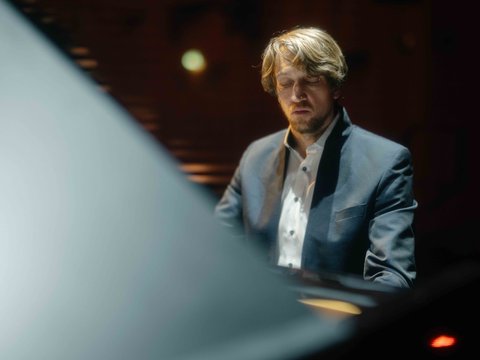 A man in a suit plays piano intently on a dimly lit stage, with soft lighting highlighting the scene.