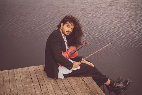 A musician with long hair sits on a wooden dock by the water, holding a violin. He's wearing a suit.