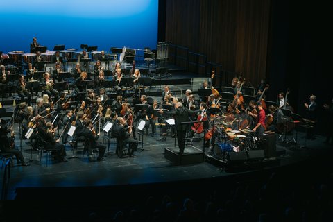 Ein Orchester spielt auf einer Bühne vor einem blauen Hintergrund.
