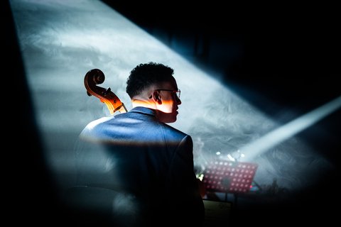 Ein Mann spielt das Violoncello in einem Konzert. Sein Rücken ist zur Kamera, und sein Kopf ist nach rechts gedreht, sodass sein Gesicht im Profil ist.