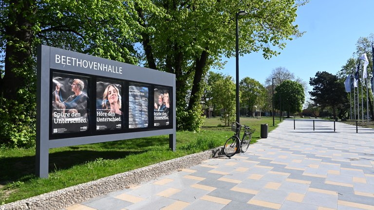 Billboard outside Beethovenhalle displaying advertisement posters. A sunny path lined with trees stretches into the distance.