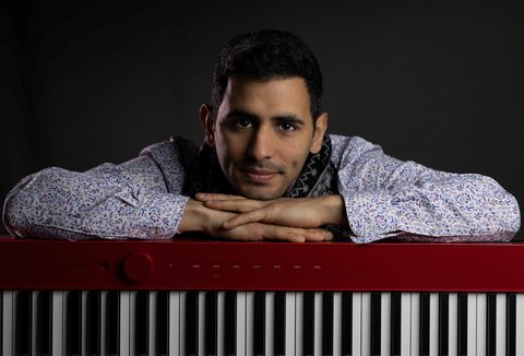A man leans on a red keyboard, smiling. He wears a patterned shirt. The background is dark.