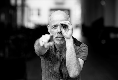 A man is standing in front of a dark background. He is wearing a patterned top. He is looking into the camera. His right hand is pointing into the camera, his left hand forms a circle around one eye.