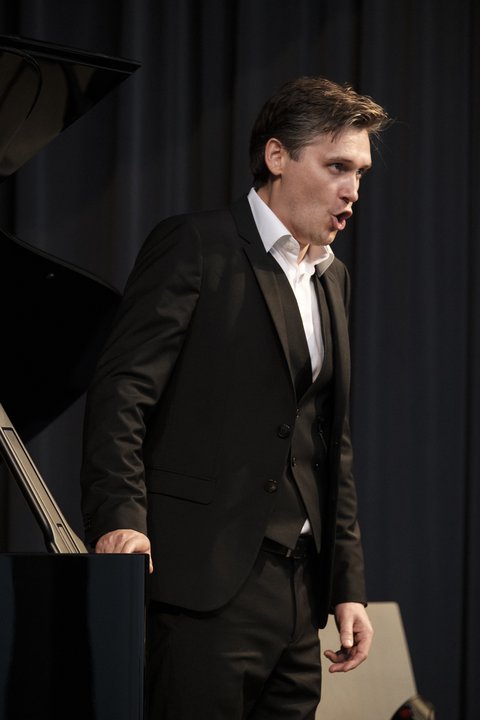 Ein Sänger steht an einem Flügel und singt. Er trägt einen schwarzen Anzug und ein weißes Hemd. Er schaut intensiv nach vorne. Der Hintergrund ist schwarz. © Niclas Weber
