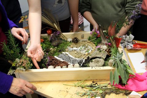 Mehrere Hände arbeiten an einem Kunstprojekt, das Naturmaterialien wie Zweige, Blätter und Steine in einer Holzkiste umfasst.