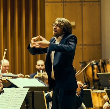 Ein Dirigent in einem Konzertsaal leitet ein Orchester, während die Musiker auf ihre Notenblätter schauen und ihre Instrumente spielen.
