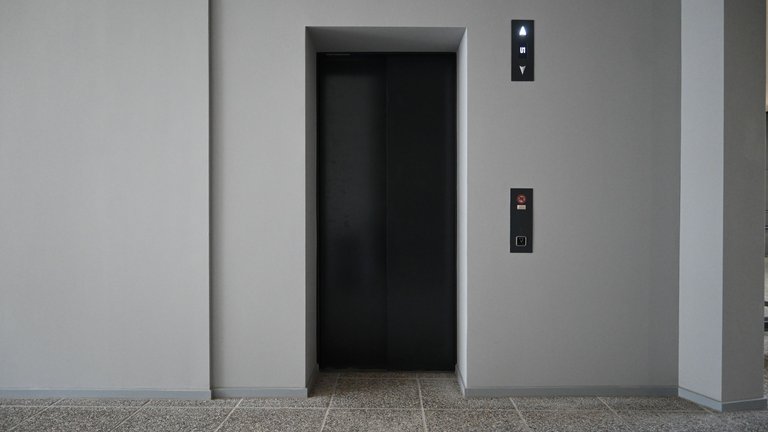 A modern elevator in a building. The open doors reveal a minimalist design with gray walls and a stone-like floor.