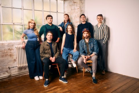 A group of eight people pose in a bright room, some seated and some standing, next to a large window.